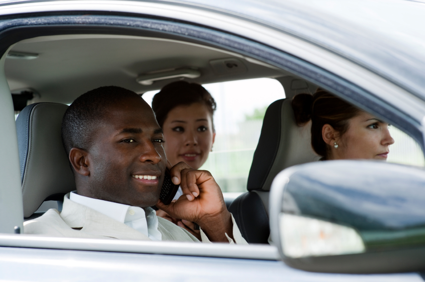 Three people sitting inside a car. A man in a light suit is in the passenger's seat, smiling while talking on a cellphone. Two women are seated behind him, looking forward. The car window is open.